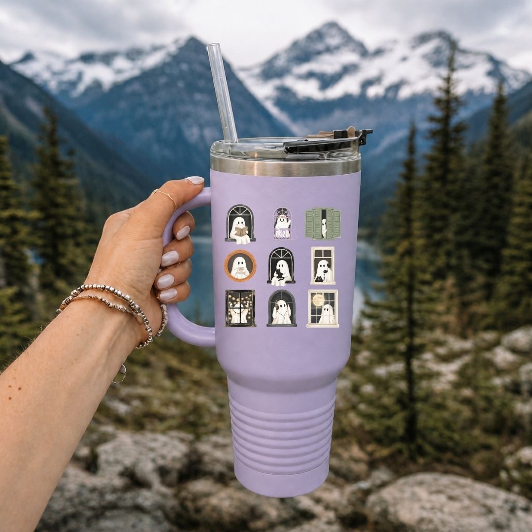 A lavender purple insulated tumbler with a clear straw and lid, held by a hand against a mountain and forest background. The tumbler features a graphic print of nine ghost-themed tombstone illustrations.