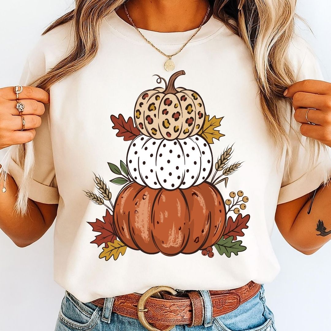 A woman wearing a cream-colored t-shirt with a graphic of three stacked pumpkins in autumn colors, paired with blue jeans and a brown leather belt.