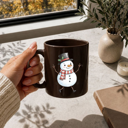 A hand holds a dark brown ceramic mug featuring a cartoon snowman wearing a black top hat and a red-and-white striped scarf. The mug is shown in a warm, sunlit indoor setting on a light stone surface near a window, with a beige vase, candle, and notebook in the background.