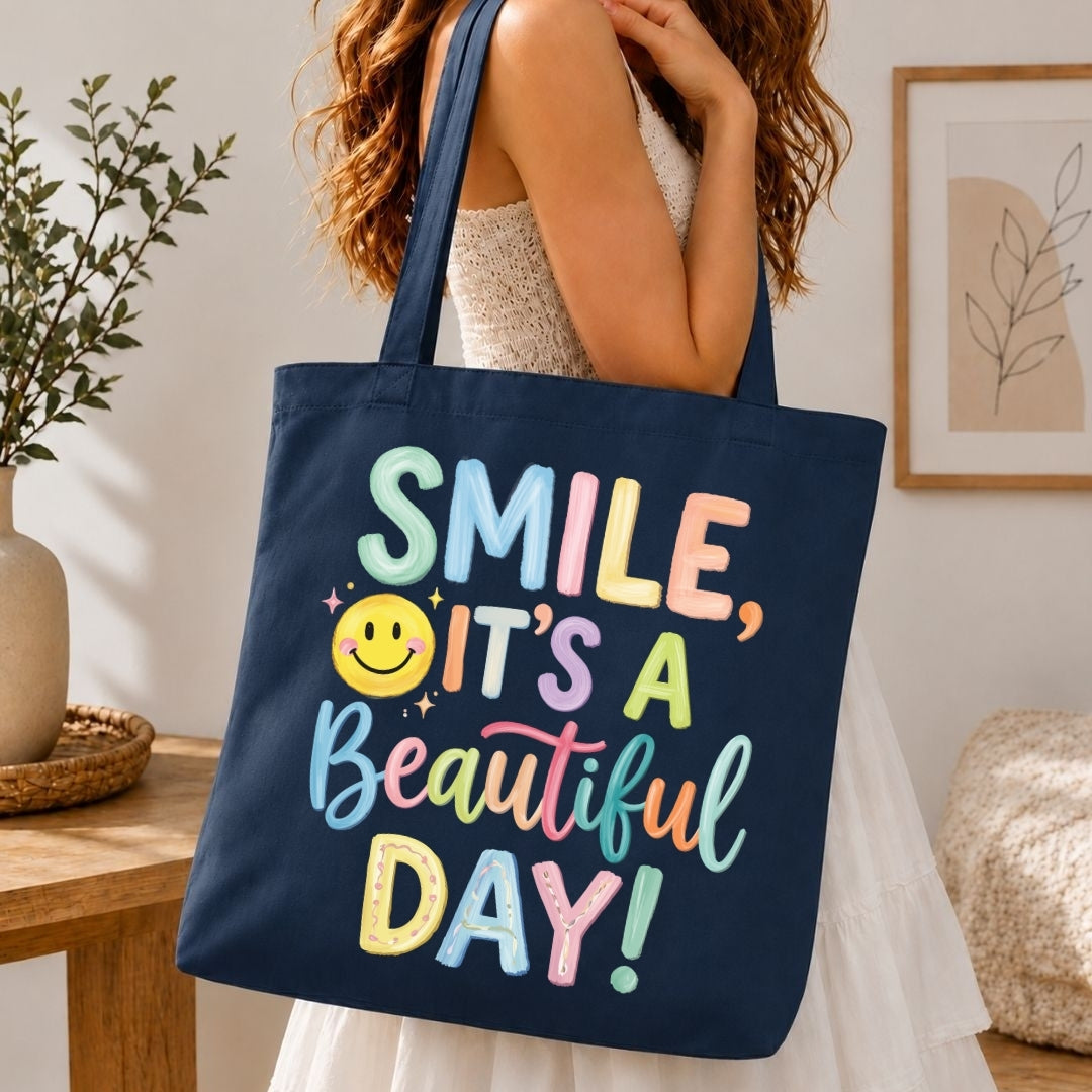 A navy blue canvas tote bag with colorful text reading 'SMILE, IT'S A Beautiful DAY!' and a yellow smiley face, carried by a woman in a white dress in a bright indoor setting.