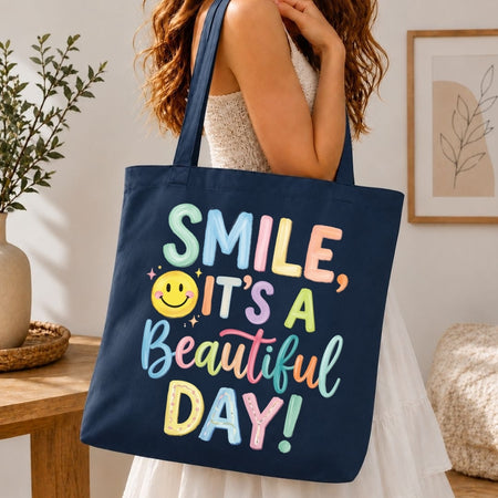 A navy blue canvas tote bag with colorful text reading 'SMILE, IT'S A Beautiful DAY!' and a yellow smiley face, carried by a woman in a white dress in a bright indoor setting.