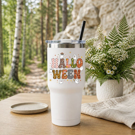 A white insulated tumbler with a clear lid and black straw sits on a wooden table outdoors. The tumbler is printed with the word "HALLOWEEN" in patterned letters and small ghost illustrations.