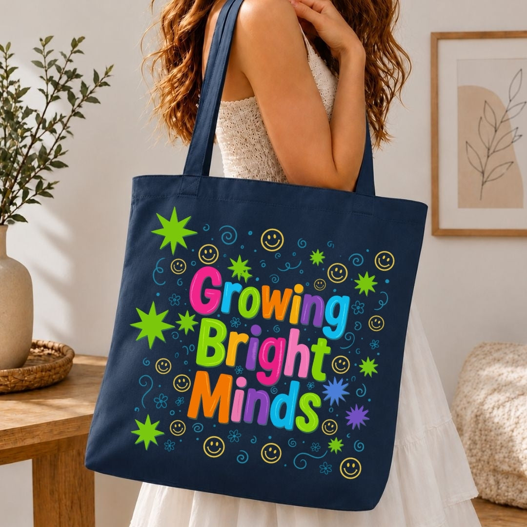 A navy blue canvas tote bag with the phrase 'Growing Bright Minds' in colorful letters, surrounded by smiley faces and starburst shapes, carried by a woman in a white dress in a brightly lit room.