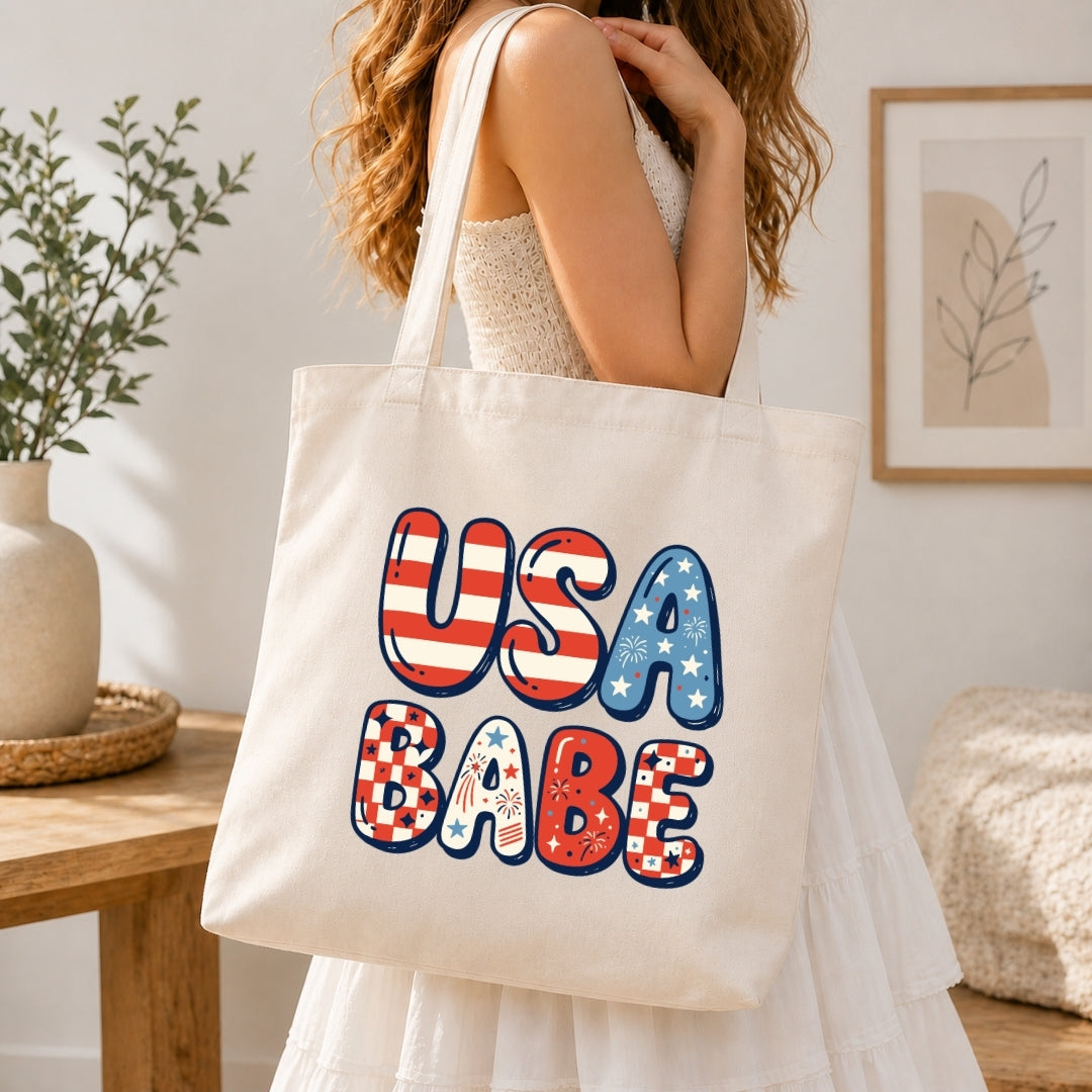 A woman in a white dress carrying a large cream-colored canvas tote bag over her shoulder. The center of the tote bag showcases the large bubble-lettered "USA BABE" graphic decorated with stars, stripes, and checkers.