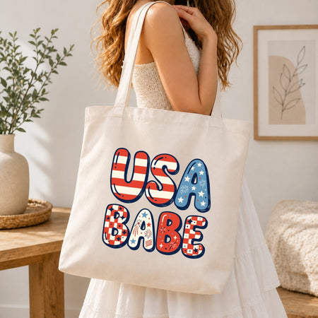 A woman in a white dress carrying a large cream-colored canvas tote bag over her shoulder. The center of the tote bag showcases the large bubble-lettered "USA BABE" graphic decorated with stars, stripes, and checkers.