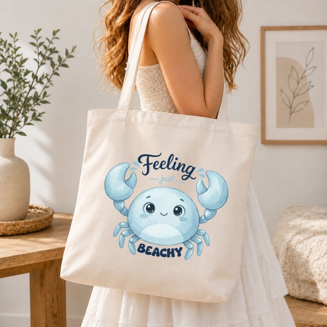 A woman holding a cream-colored tote bag printed with a blue cartoon crab graphic and the text "Feeling just BEACHY".