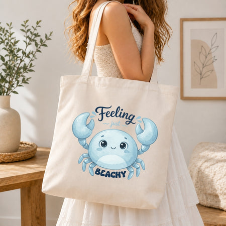 A woman holding a cream-colored tote bag printed with a blue cartoon crab graphic and the text "Feeling just BEACHY".