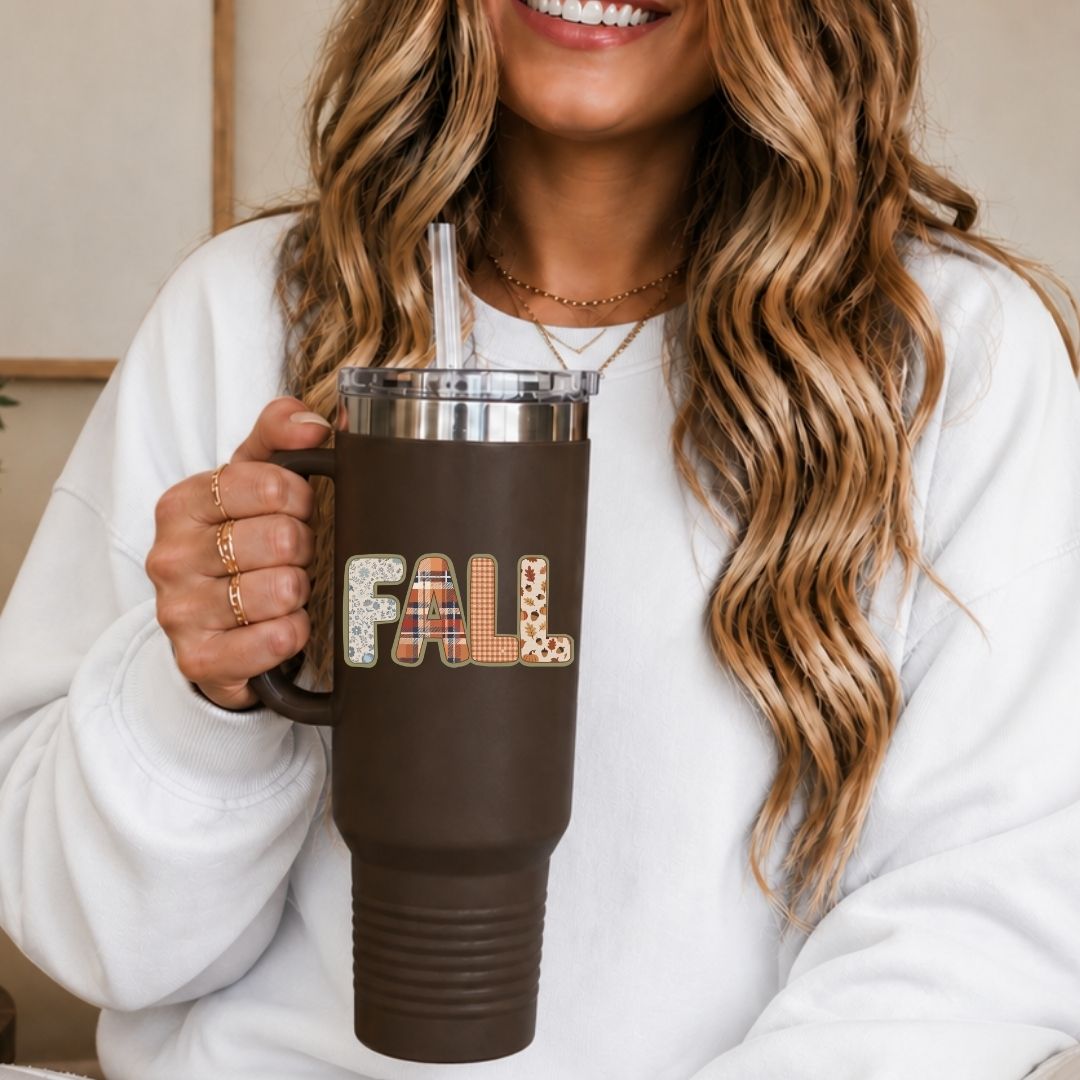 A woman in a white sweatshirt holds a brown insulated tumbler with a clear lid and straw. The tumbler has a dark brown sleeve with the word "FALL" in patterned letters.
