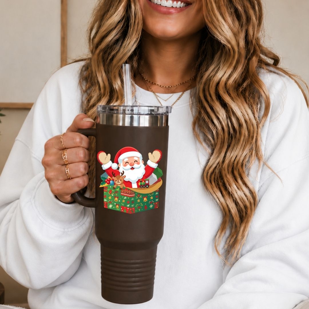 A woman in a white sweatshirt holds a dark brown insulated tumbler with a clear lid and straw. The tumbler features a colorful graphic of Santa Claus sitting in a sleigh decorated with Christmas trees and ornaments.