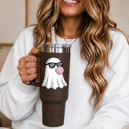 A woman in a white sweatshirt holds a brown insulated tumbler with a clear lid and straw. The tumbler has a printed graphic of a white ghost wearing black sunglasses and blowing a pink bubblegum bubble.