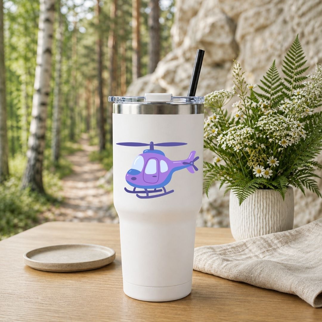 A white insulated tumbler with a clear lid and black straw sits on a wooden table outdoors. The tumbler is printed with a purple and blue helicopter graphic.