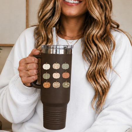 A woman in a white sweatshirt holds a dark brown insulated tumbler with a handle, clear lid, straw, and a printed pumpkin pattern in autumn colors.