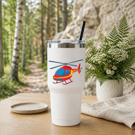 A white insulated travel tumbler with a clear lid and black straw, featuring a colorful helicopter graphic, set on a wooden table with a forest and stone wall background.