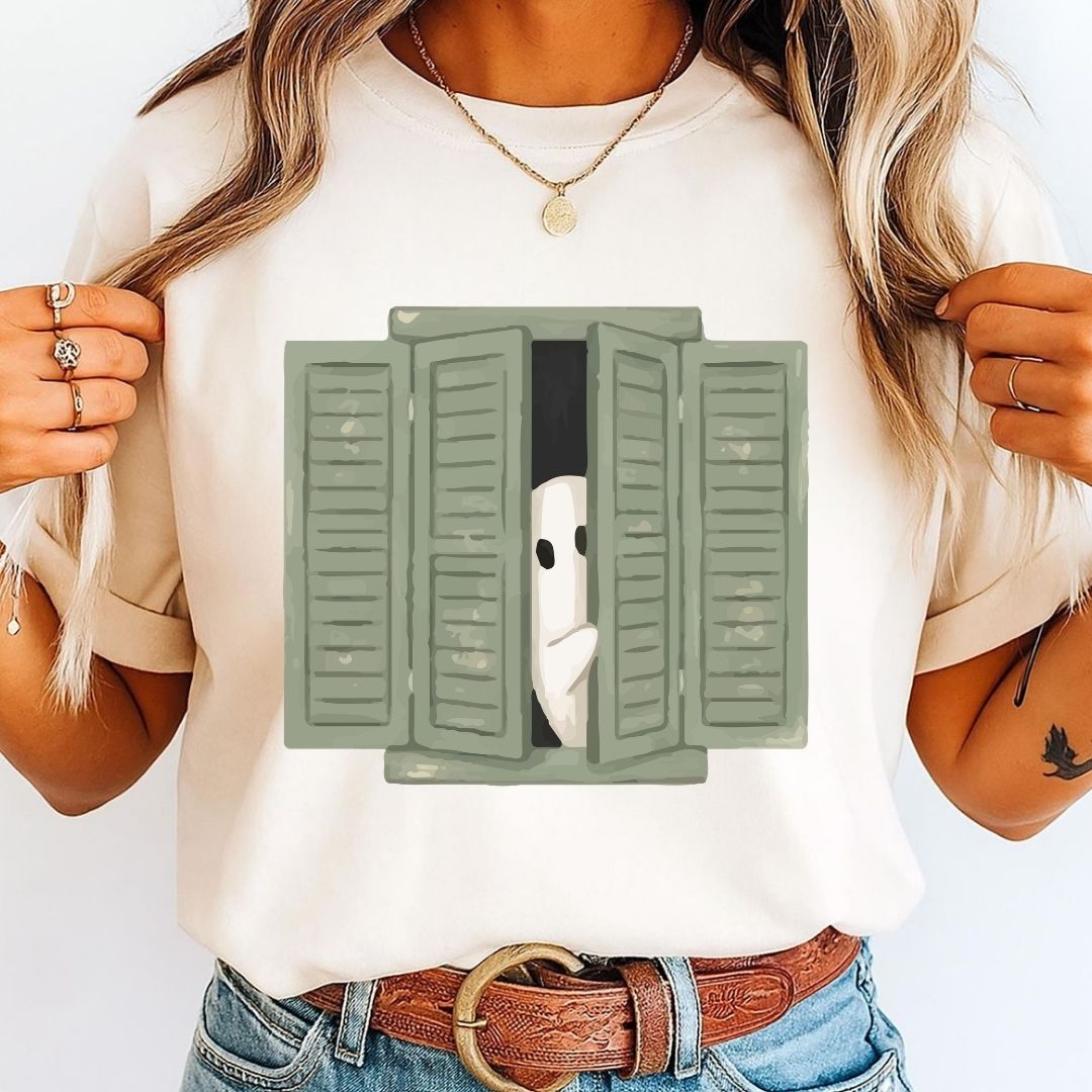 A woman wearing a cream-colored t-shirt with a graphic of a white ghost peeking out from behind green shutters, paired with blue jeans and a brown belt.