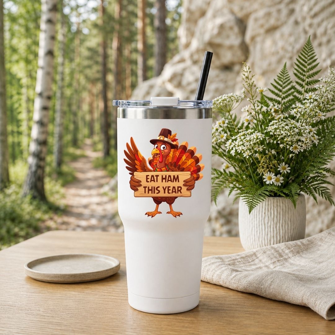 A white insulated tumbler with a clear lid and black straw stands on a wooden table outdoors. The tumbler has a printed cartoon turkey holding a sign that reads “EAT HAM THIS YEAR.