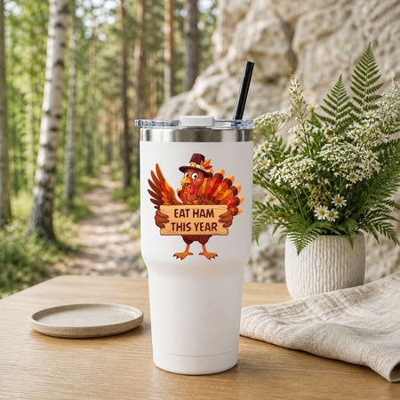 A white insulated tumbler with a clear lid and black straw stands on a wooden table outdoors. The tumbler has a printed cartoon turkey holding a sign that reads “EAT HAM THIS YEAR.