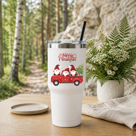 A white insulated tumbler with a clear lid and black straw stands on a wooden table in a forest setting. The tumbler is decorated with a red truck carrying three Santa Clauses and the words 'Merry Christmas.