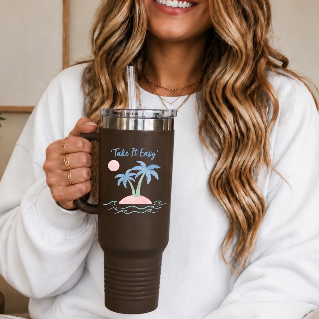 A woman in a white sweatshirt holds a dark brown insulated tumbler with a clear lid and straw. The tumbler has a tropical island graphic with palm trees, blue waves, a pink sun, and the words 'Take It Easy'.