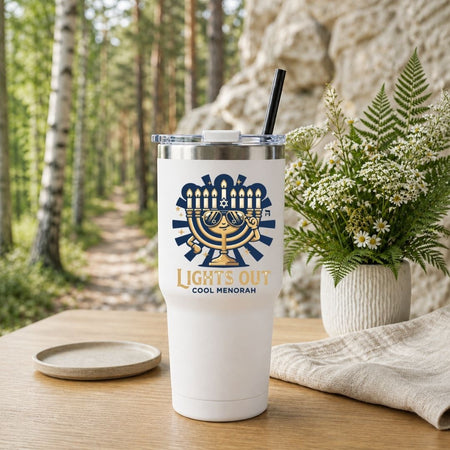 A white insulated tumbler with a clear lid and black straw sits on a wooden table outdoors. The tumbler has a navy and gold menorah graphic with the words 'LIGHTS OUT COOL MENORAH.