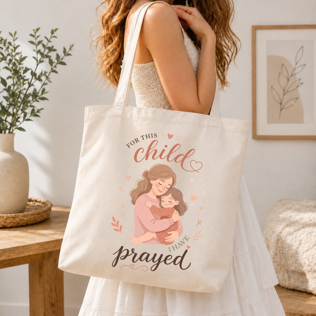 A woman holding a cream-colored tote bag printed with a mother-and-child illustration and the text "For this child I have prayed."