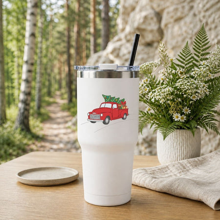 A white insulated tumbler with a clear lid and black straw sits on a wooden table outdoors. The tumbler is printed with a red vintage truck carrying a Christmas tree.