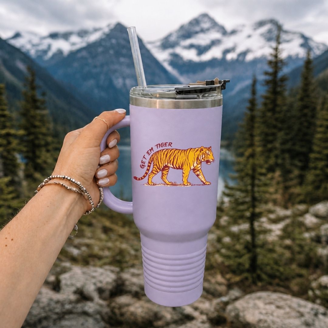 A lavender insulated travel tumbler with a handle, clear straw, and lid, held by a hand against a mountain and forest background. The tumbler features a graphic of an orange tiger with the text 'LET IT RAIN TIGER'.