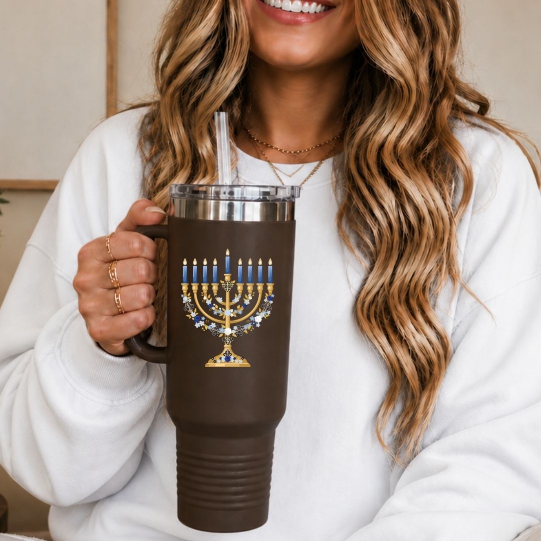 A woman in a white sweatshirt holds a brown insulated tumbler with a clear lid and straw. The tumbler features a gold and blue menorah graphic with candle details.