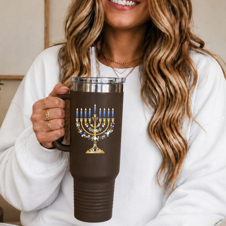 A woman in a white sweatshirt holds a brown insulated tumbler with a clear lid and straw. The tumbler features a gold and blue menorah graphic with candle details.