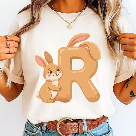 A woman holding the sides of an off-white t-shirt featuring a light brown letter R with bunny ears and a friendly cartoon rabbit hugging it.