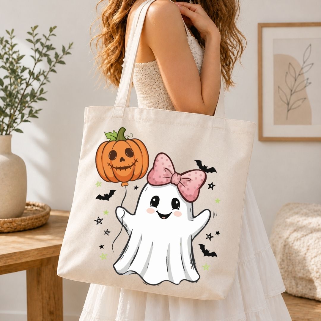 A woman wearing a white dress carries a cream canvas tote bag decorated with a cute cartoon ghost, a jack-o'-lantern pumpkin, pink bow, and small bats, set against a warm, softly lit indoor background.
