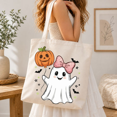 A woman wearing a white dress carries a cream canvas tote bag decorated with a cute cartoon ghost, a jack-o'-lantern pumpkin, pink bow, and small bats, set against a warm, softly lit indoor background.