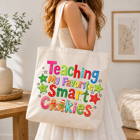 A woman in a white dress carries a cream canvas tote bag with colorful text reading 'Teaching My Favorite Smart Cookies' and decorated stars and cookies. The scene is set in a bright, modern room with a wooden table, potted plant, and framed wall art.