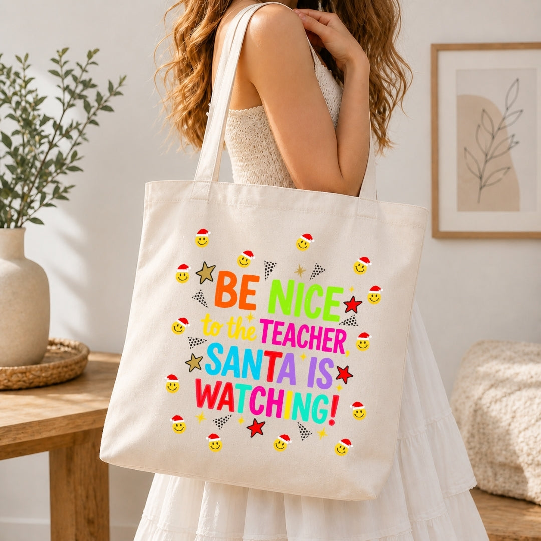 A woman in a white dress carries a cream canvas tote bag with colorful text reading 'BE NICE TO THE TEACHER SANTA IS WATCHING!' and small Santa-themed smiley faces and stars, shown in a bright indoor room.