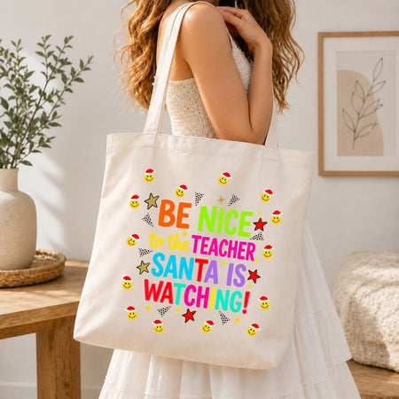 A woman in a white dress carries a cream canvas tote bag with colorful text reading 'BE NICE TO THE TEACHER SANTA IS WATCHING!' and small Santa-themed smiley faces and stars, shown in a bright indoor room.