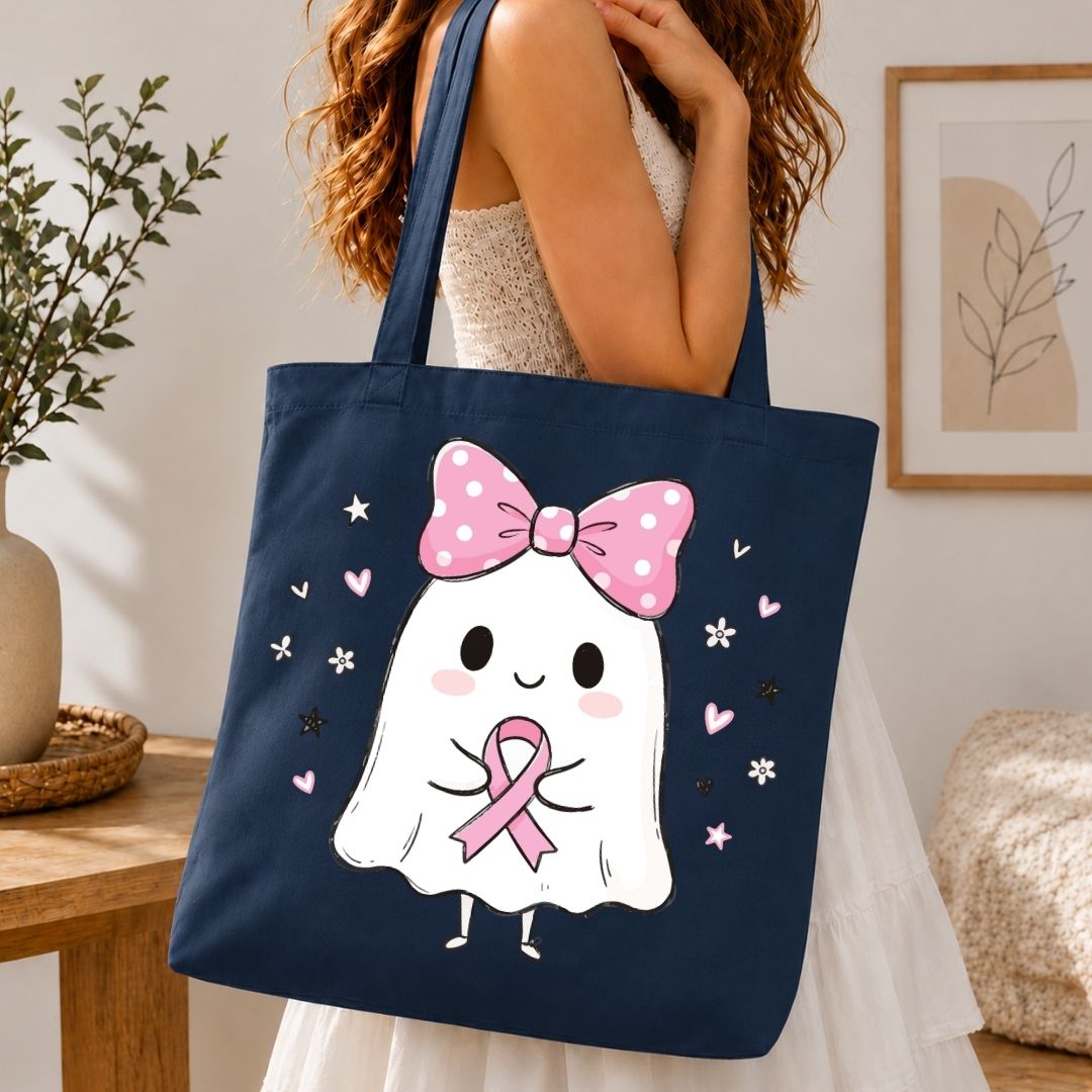 A navy blue canvas tote bag with a cute cartoon ghost graphic wearing a pink polka-dot bow and holding a pink ribbon, carried by a woman in a white dress in a cozy indoor setting.