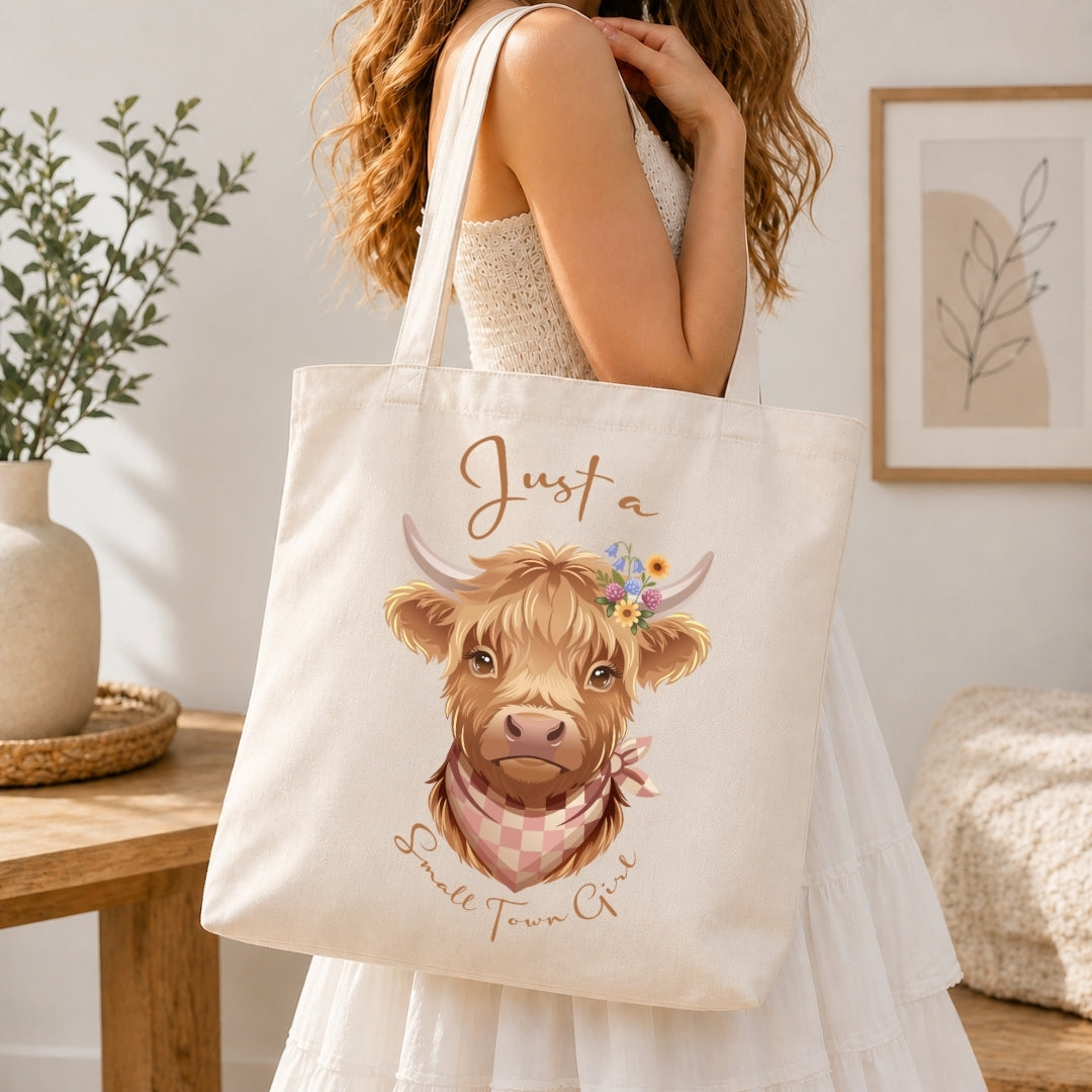 A woman in a white dress carries a cream canvas tote bag featuring a cute illustration of a Scottish highland cow with the text 'Just a Small Town Girl' in brown script.