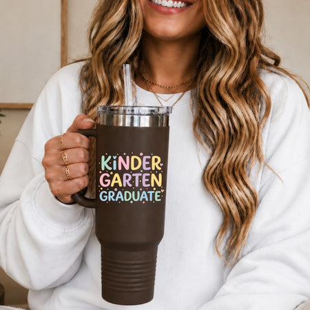 A woman in a white sweatshirt holds a dark brown insulated tumbler with a clear lid and straw. The tumbler has colorful text reading 'KINDER GARTEN GRADUATE' in pastel rainbow letters.