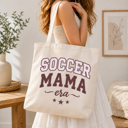 Woman in a white dress carrying a canvas tote bag printed with a maroon and pink vintage "Soccer Mama Era" sports design over her shoulder.