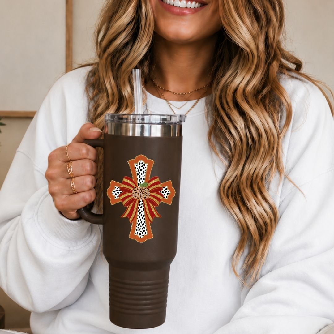 A woman in a white sweatshirt holds a brown insulated tumbler with a clear lid and straw. The tumbler features a large orange and white cross design with a small green floral accent at the center.