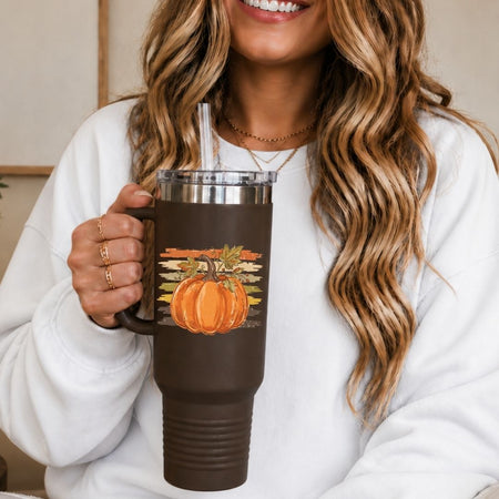 A woman in a white sweatshirt holds a dark brown insulated tumbler with a handle, clear lid, and straw. The tumbler features a printed autumn design with an orange pumpkin and colorful leaves.