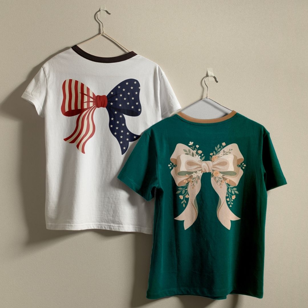 Two t-shirts hanging side-by-side against a wall: a white ringer tee featuring a bow patterned with American flag stars and stripes, and a dark green tee featuring an off-white bow adorned with delicate floral branches.