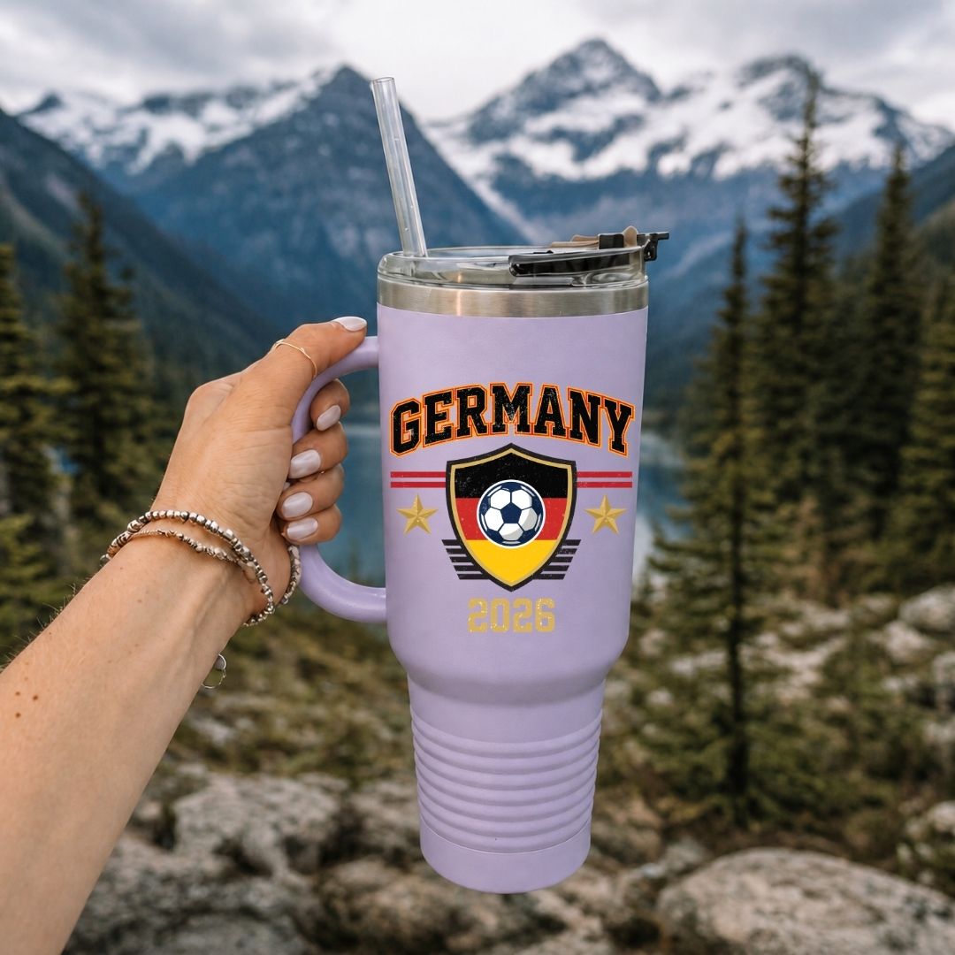 A close up of a person holding a large 40oz lavender purple travel tumbler with a handle, featuring a retro Germany soccer graphic with mountains and an alpine lake in the background.