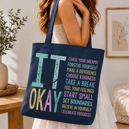 A navy blue canvas tote bag with colorful text reading 'IT is OKAY' and a motivational quote, carried by a woman in a white dress in a bright indoor setting.