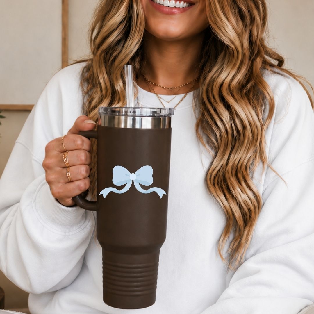 A woman in a white sweatshirt holds a dark brown insulated tumbler with a clear lid and straw. The tumbler has a light blue bow graphic on the front.
