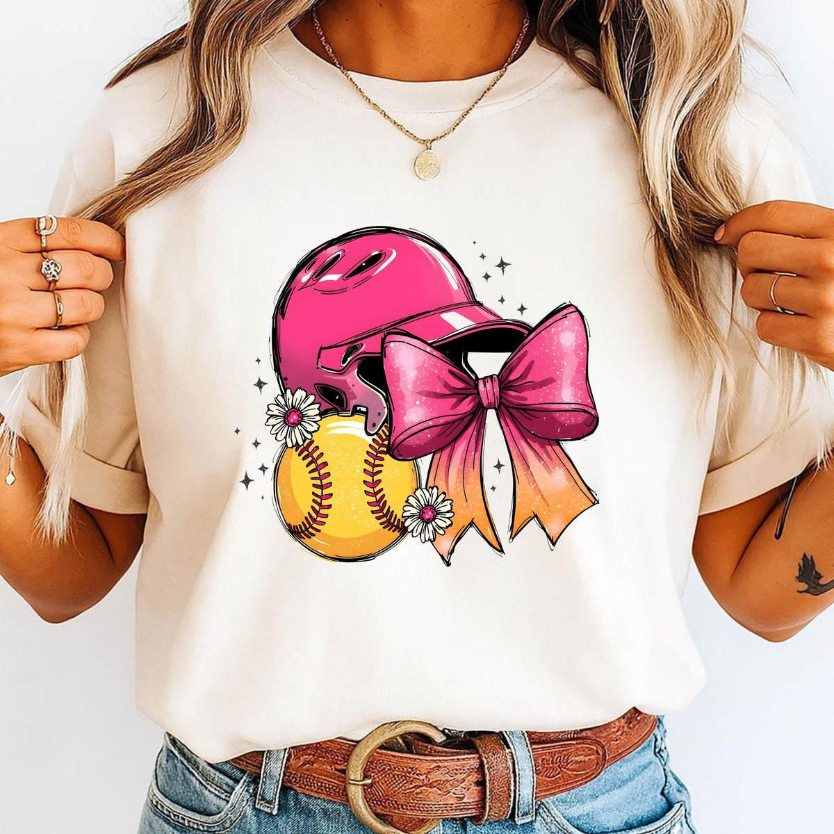A woman wearing a cream-colored t-shirt with a graphic of a pink softball helmet, a yellow softball, and a large pink bow, paired with blue jeans and a brown belt.