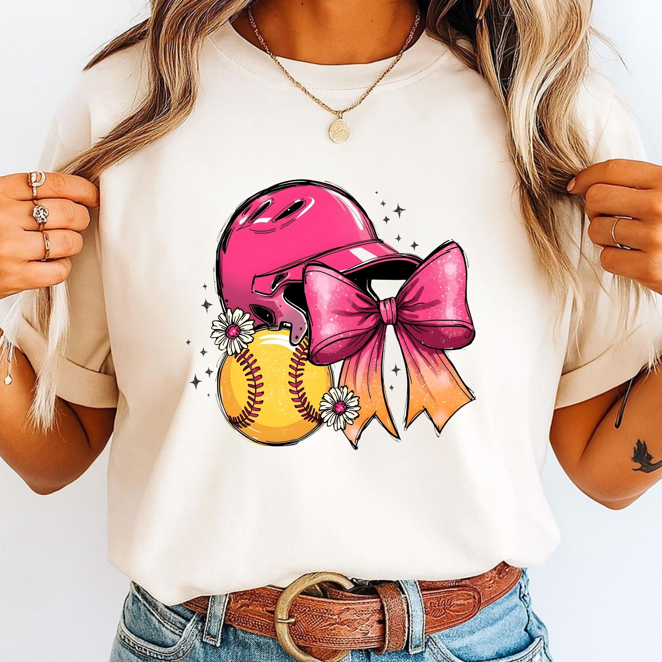 A woman wearing a cream-colored t-shirt with a graphic of a pink softball helmet, a yellow softball, and a large pink bow, paired with blue jeans and a brown belt.