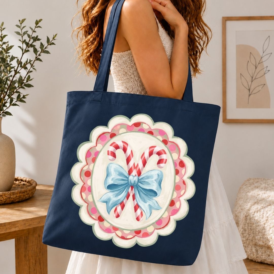 A navy blue canvas tote bag with a large holiday graphic of two candy canes in a blue bow on a scalloped pink and white background, carried by a woman in a white dress in a cozy indoor setting.