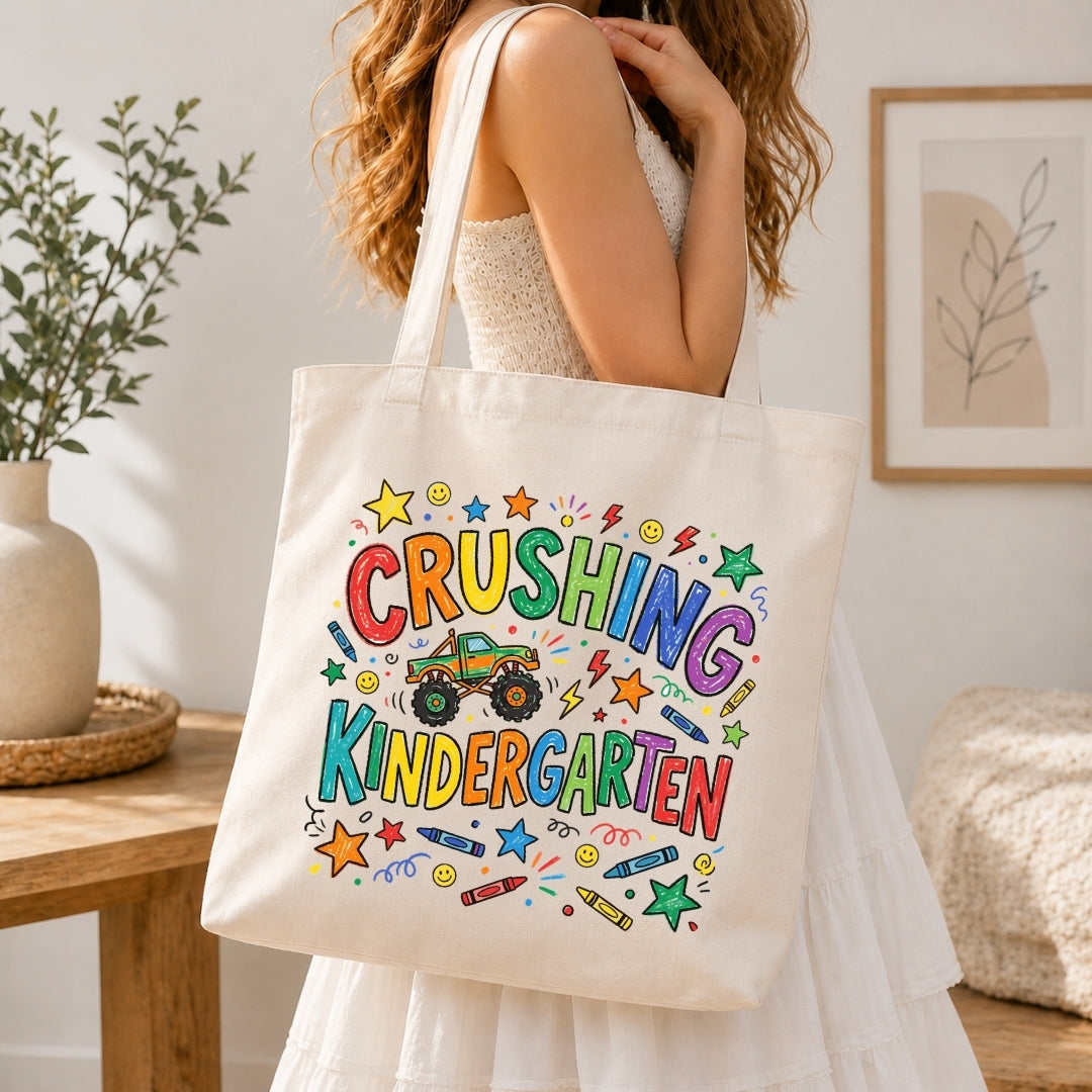 A woman in a white dress carries a cream canvas tote bag with a colorful 'CRUSHING KINDERGARTEN' graphic featuring a monster truck and crayons, set in a bright, modern room.