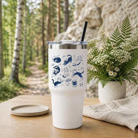 A white insulated tumbler with a clear lid and black straw, decorated with blue whimsical duck illustrations, set on a wooden table outdoors with a forest path, rock wall, and potted fern in the background.