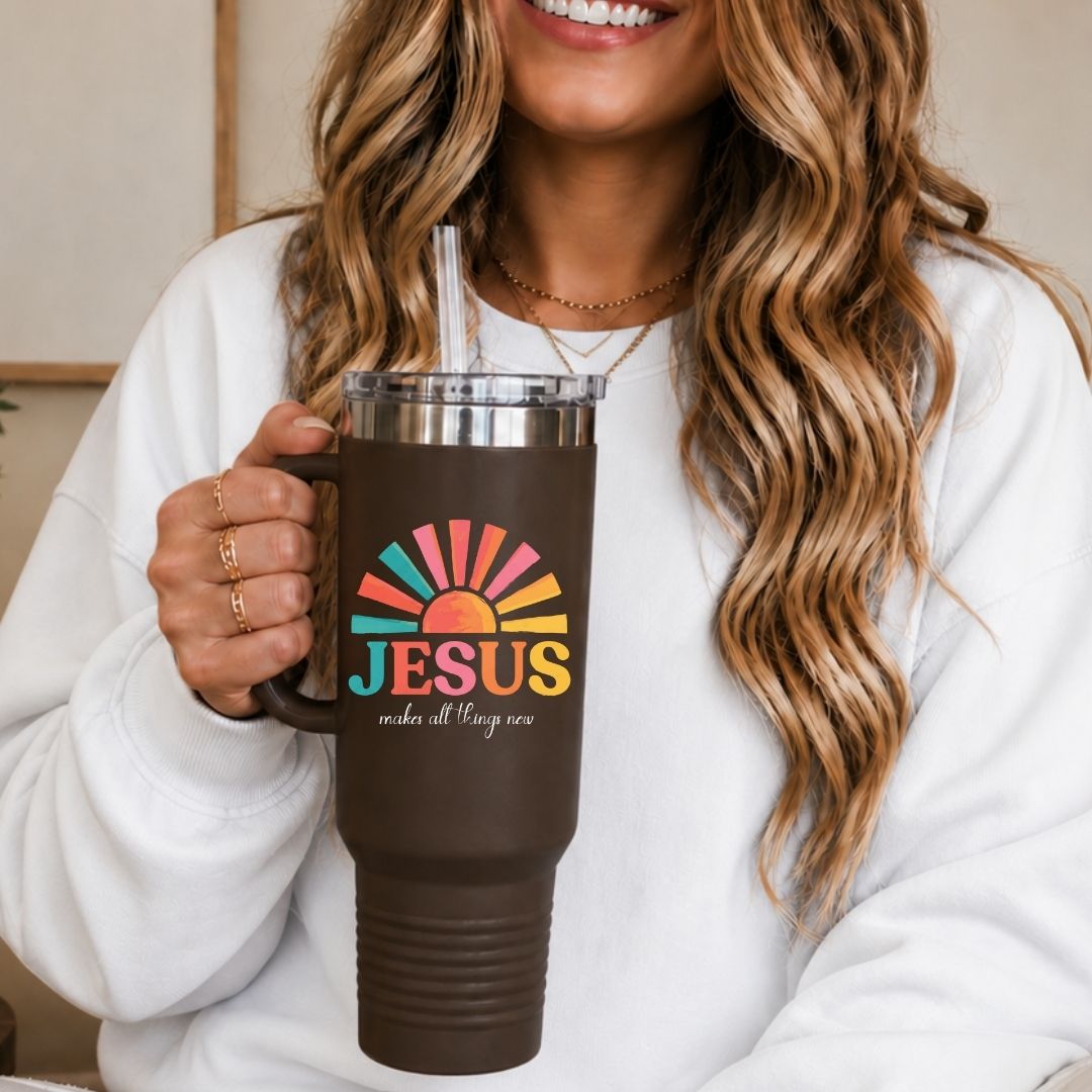 A woman in a white sweatshirt holds a dark brown insulated tumbler with a clear lid and straw. The tumbler features a colorful sunburst graphic and the text 'JESUS makes all things new'.